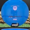 Blue disc golf disc with Discmania and Armory Disc Golf branding on a grass and log background.